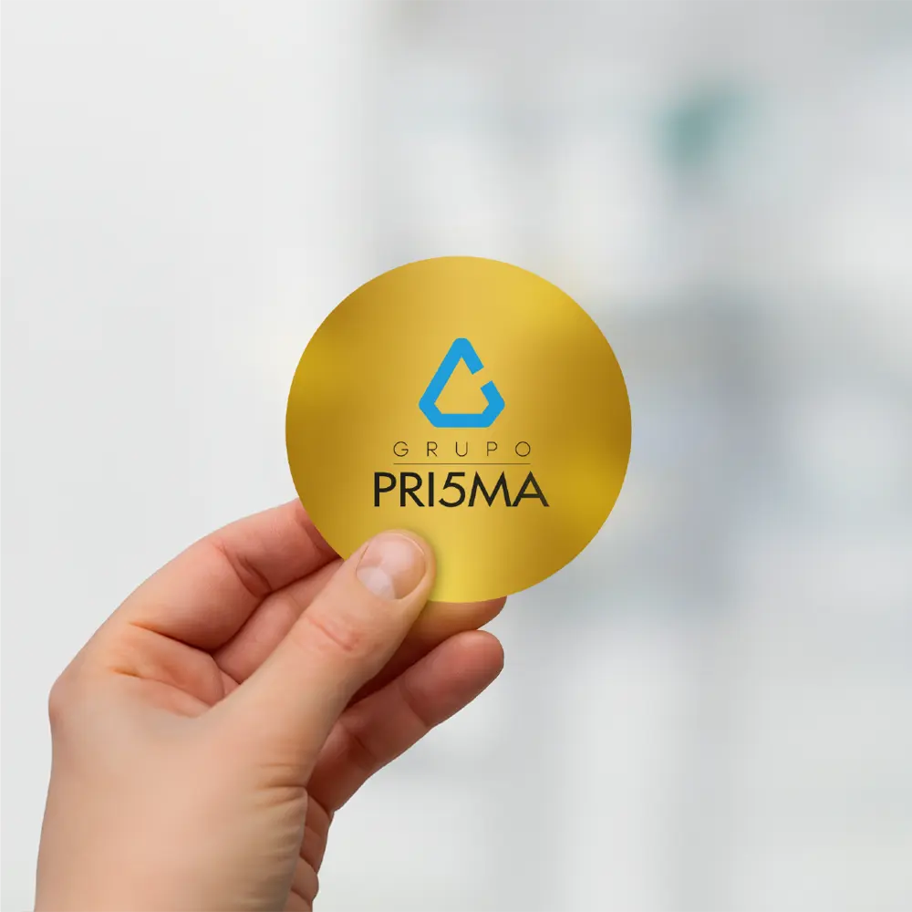 Mano sosteniendo un sticker dorado con el logo de Grupo PRI5MA